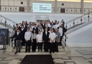 Na zdjęciu widoczna jest grupa uczniów wraz z opiekunami podczas wizyty edukacyjnej w gmachu Sejmu Rzeczypospolitej Polskiej. Uczestnicy pozują do wspólnego zdjęcia na reprezentacyjnych schodach, a w tle znajduje się ekran z napisem „Rok Ignacego Daszyńskiego 2026”, nawiązujący do patrona roku.