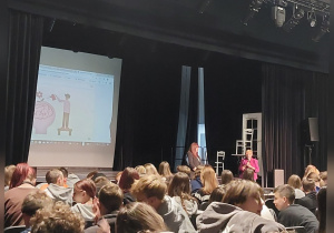 Uczniów z widowni oglądają dwie osoby prowadzące edukacyjny spektakl.