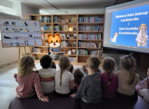 Dzieci zgromadzone w bibliotece, biorą udział w zajęciach poświęconych tematyce zwierząt. Przed nimi znajduje się prowadzący, a w tle widoczny jest ekran z informacją o Światowym Dniu Zwierz