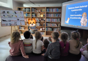 Dzieci zgromadzone w bibliotece, biorą udział w zajęciach poświęconych tematyce zwierząt. Przed nimi znajduje się prowadzący, a w tle widoczny jest ekran z informacją o Światowym Dniu Zwierząt oraz Dniu Kundelka