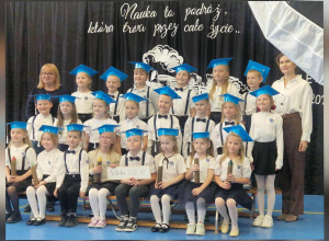 Uroczyste ślubowanie pierwszoklasistów. Dzieci ubrane są odświętnie i ustawione w trzech rzędach na tle dekoracji z motywem edukacyjnym.