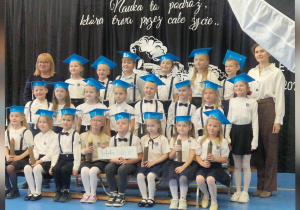 Uroczyste ślubowanie pierwszoklasistów. Dzieci ubrane są odświętnie i ustawione w trzech rzędach na tle dekoracji z motywem edukacyjnym.