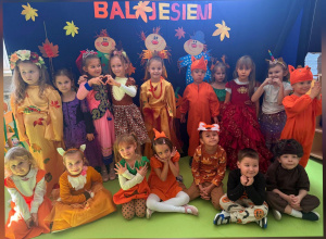 Przedszkolaki w barwnych, jesiennych strojach prezentują się na tle dekoracji przygotowanej z okazji Balu Jesieni