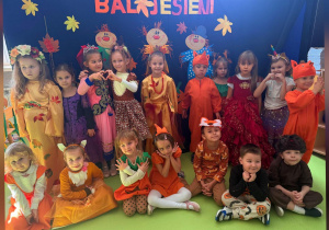 Przedszkolaki w barwnych, jesiennych strojach prezentują się na tle dekoracji przygotowanej z okazji Balu Jesieni