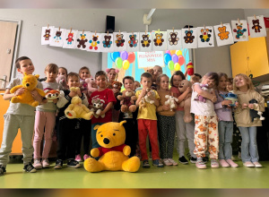Dzieci świętują Dzień Pluszowego Misia w kolorowo udekorowanej sali, trzymając swoje ulubione pluszaki – wśród nich króluje duży Kubuś Puchatek.