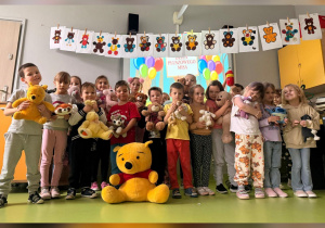 Dzieci świętują Dzień Pluszowego Misia w kolorowo udekorowanej sali, trzymając swoje ulubione pluszaki – wśród nich króluje duży Kubuś Puchatek.