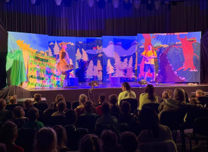 Przedszkolaki na widowni podczas przedstawienia. W tle kolorowa dekoracja.