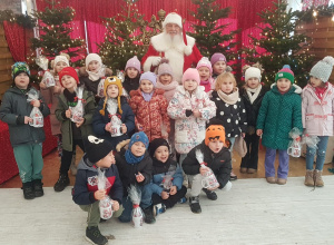 Przedszkolaki z Mikołajem w zimowej, świątecznej scenerii pełnej prezentów i światełek.