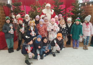 Przedszkolaki z Mikołajem w zimowej, świątecznej scenerii pełnej prezentów i światełek.