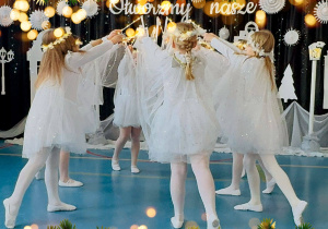 Dziewczynki tańczą ubrane w białe sukienki, a w dłoniach trzymają różdżki.