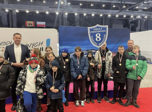 Grupa uczniów z medalami i opiekunami stoi w hali sportowej z tarczą swojej szkoły.