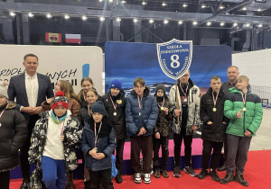 Grupa uczniów z medalami i opiekunami stoi w hali sportowej z tarczą swojej szkoły.