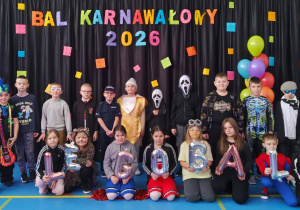 Dzieci w kolorowych przebraniach pozujących przed dekoracją z napisem „Bal Karnawałowy 2026” i motywem LEGO.