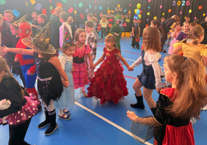 Dzieci w kolorowych strojach trzymają się za ręce i tańczą podczas balu karnawałowego.