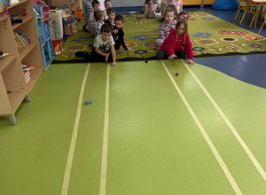 Przedszkolaki bawią się samochodzikami, przesuwając je po podłodze w kierunku mety.