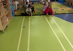 Przedszkolaki bawią się samochodzikami, przesuwając je po podłodze w kierunku mety.