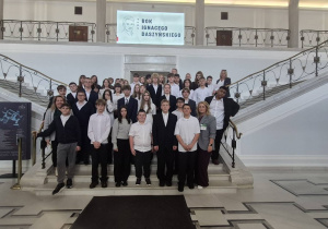 Na zdjęciu widoczna jest grupa uczniów wraz z opiekunami podczas wizyty edukacyjnej w gmachu Sejmu Rzeczypospolitej Polskiej. Uczestnicy pozują do wspólnego zdjęcia na reprezentacyjnych schodach, a w tle znajduje się ekran z napisem „Rok Ignacego Daszyńskiego 2026”, nawiązujący do patrona roku.
